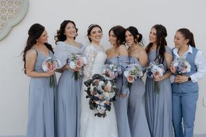 Bridal party wearing dusty blue dresses pose together while holding purple and white bouquets and laughing joyfully.