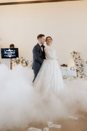 Wedding couple embracing in an elegant white room with fog effects and floral decorations.