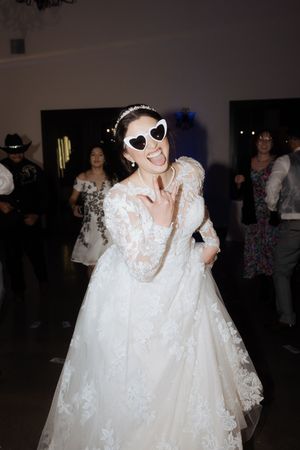 Bride in white lace wedding dress and sunglasses poses playfully indoors at reception venue.