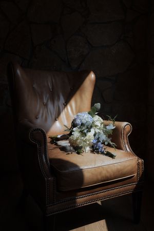 Faded hydrangea flowers rest on a vintage brown leather armchair in dramatic low-key lighting.