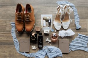 Wedding accessories and shoes laid flat on wooden floor with brown leather shoes and white lace heels arranged neatly.