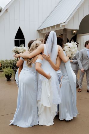A bride and bridesmaids in pale blue dresses share an intimate embrace outside a white chapel.