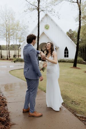 Couple shares a sweet first look moment outside a white chapel with natural woodland backdrop.