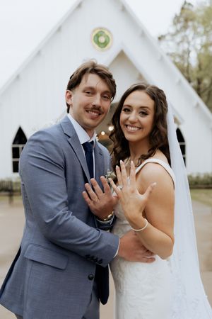 Happy newlyweds display their wedding rings with joy in front of a white chapel.