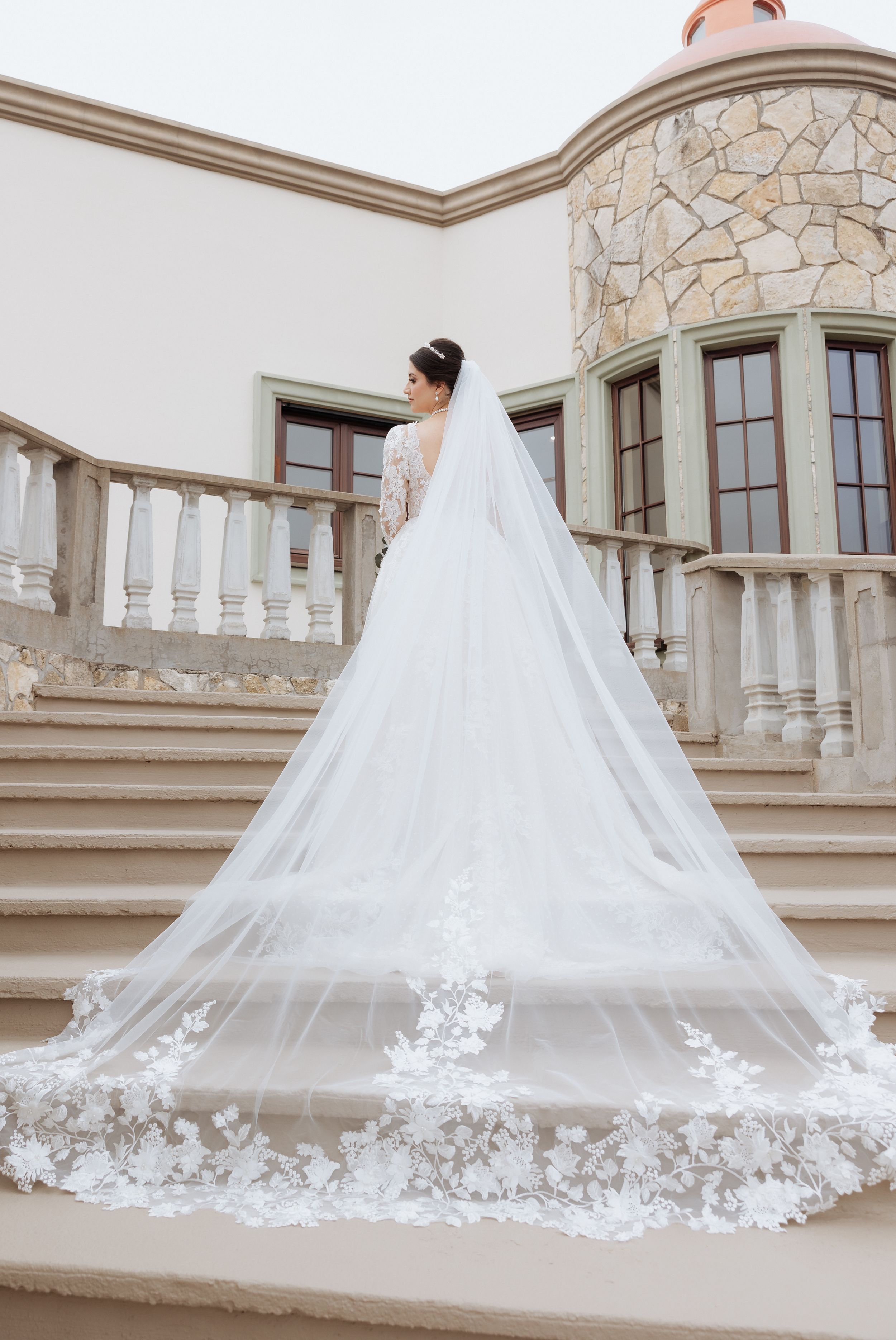 Long cathedral veil with delicate lace appliques trailing behind bride on stone balcony.