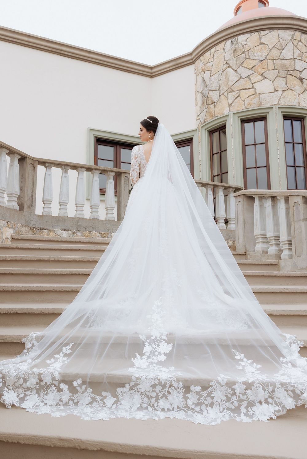 Long cathedral veil with delicate lace appliques trailing behind bride on stone balcony.