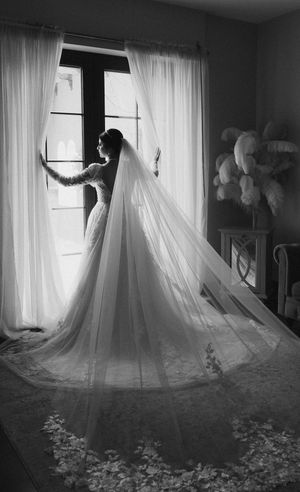 A silhouetted bride in a flowing veil stands by a bright window in a dramatic black and white photograph.