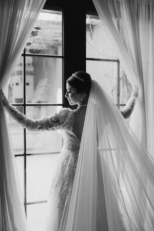 Dramatic black and white silhouette of bride in lace dress and veil standing by window with sheer curtains.