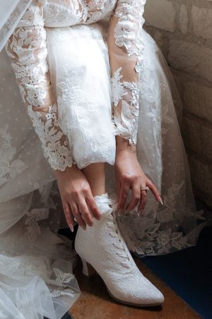 White lace wedding boots and dress details shown as bride adjusts footwear in an elegant bridal preparation moment.