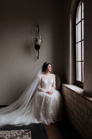 Bride in long-sleeved lace wedding gown sitting by arched window in moody church setting with flowing veil.