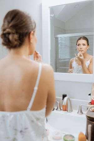 Someone applies lip product while looking in a bathroom mirror reflection in a white silk robe and spaghetti strap top.