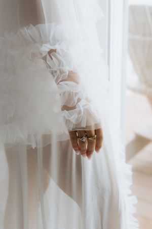 Close-up of delicate lace sleeve and engagement ring on bride's hand against white wedding dress background.