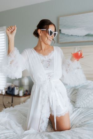 An energetic figure in a white lace robe and sunglasses dances on a bed while holding a pink cocktail in hand.