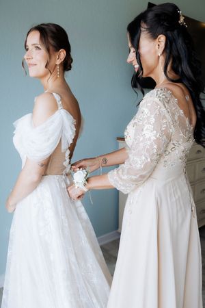 Two women in elegant white wedding dresses, one helping the other with final adjustments before the ceremony.