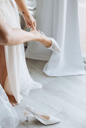 Close-up view of hands helping put on white bridal heels with lace details on a light floor.