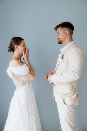A couple in matching white wedding attire share an emotional first look moment against a gray backdrop.