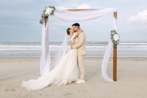 A romantic beach wedding ceremony with soft white drapery and palm trees set against the ocean waves at sunset.
