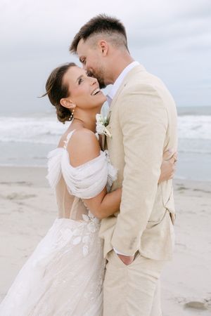 A romantic beach wedding couple embraces on a cloudy day, with the bride in white and groom in beige attire.