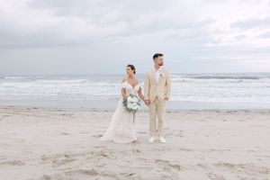 Couple in light beige and white attire walking along a serene beach with gentle waves in the background.