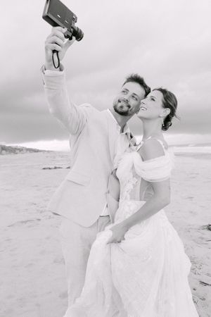 A couple in wedding attire taking a selfie together on a beach during an overcast day.