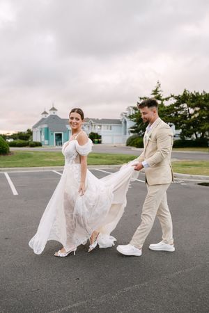 A couple in wedding attire walking in a parking lot with a white church and cloudy sky in the background.
