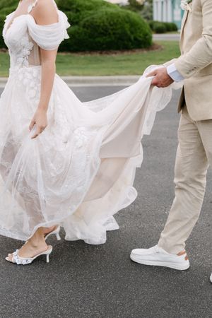 Close-up view of bride in lace dress with kitten heel shoes and groom in beige suit with white sneakers on pavement.