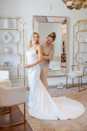 A bride in a white gown smiles as her bridesmaid helps fasten her dress in an elegant, gold-accented bridal suite.
