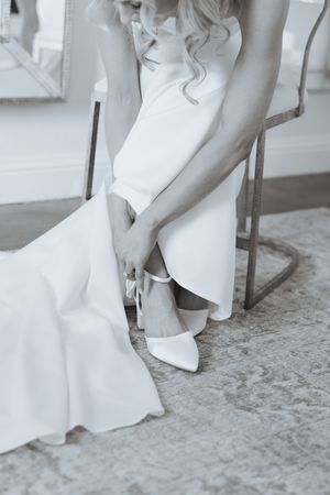 Bride in white wedding gown sitting on chair fastening elegant white heels, black and white photo.
