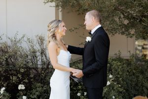 Bride in white gown and groom in black tuxedo exchanging vows outdoors, smiling joyfully during wedding ceremony.