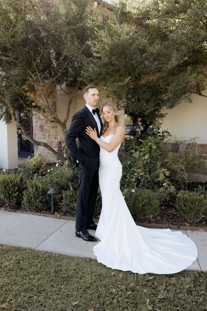 Bride in white mermaid gown embraces groom in black tuxedo outdoors near lush greenery on their wedding day.