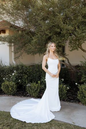 Bride in a sleek white strapless mermaid wedding gown with a train, posing outdoors near lush greenery.