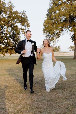 Joyful bride and groom running hand-in-hand outdoors on their wedding day, laughing under large oak trees.