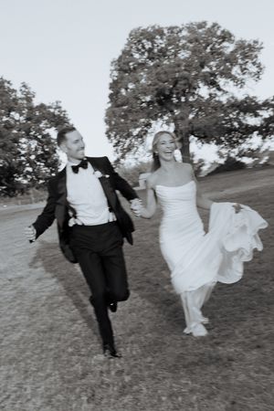 Black and white photo of a joyful bride and groom running hand in hand outdoors on their wedding day.