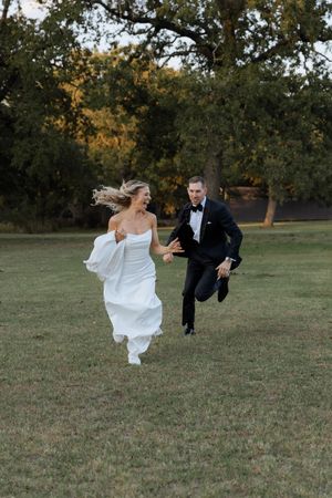 Joyful bride in white gown and groom in black tuxedo running together across a green lawn with trees in background.