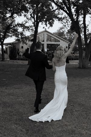 Black and white photo of newlyweds raising hands in celebration walking toward lit wedding venue.