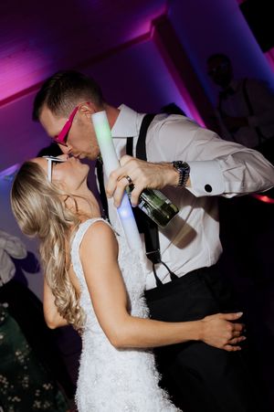 Bride and groom sharing a kiss on the dance floor, holding glow sticks and champagne bottle at their wedding reception.