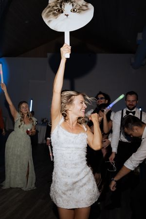 Bride in short white dress holding cat face fan while dancing at wedding reception.