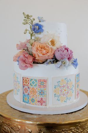 Two-tier white wedding cake with colorful mosaic tile panels and fresh pastel flowers including roses and peonies on top.