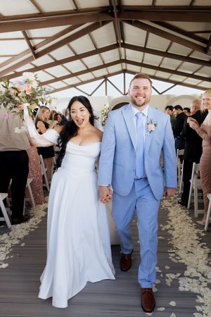 Joyful newlyweds walk down the aisle hand-in-hand, bride in white gown and groom in light blue suit.