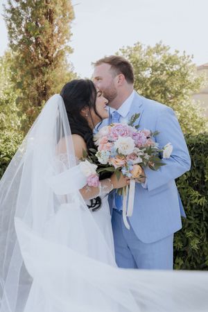 Bride and groom share a romantic kiss outdoors, bride holding a pastel bouquet, groom in light blue suit.