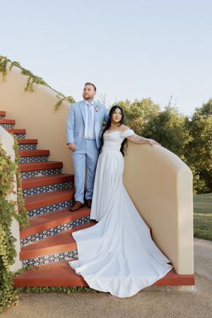 Bride in off-shoulder white gown and groom in light blue suit posing on ornate tiled staircase with ivy backdrop.