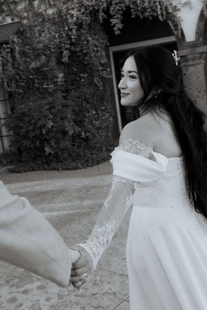 Black and white photo of dark-haired bride in lace off-shoulder wedding dress holding hands and looking back over shoulder.