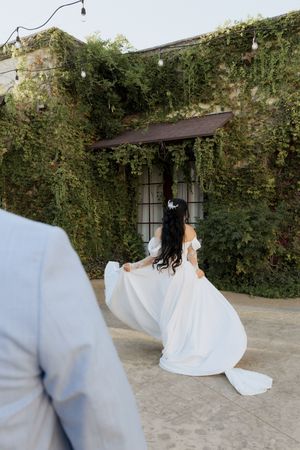 Bride in flowing off-shoulder white gown lifting her train while walking toward ivy-covered building with string lights.