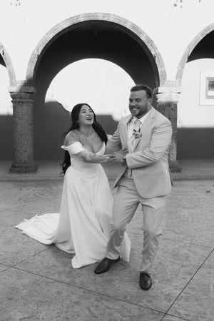 Black and white photo of laughing bride and smiling groom dancing joyfully under stone arches at their wedding.