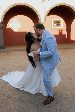 Bride and groom share a passionate dip kiss in front of arched adobe walls at golden hour.