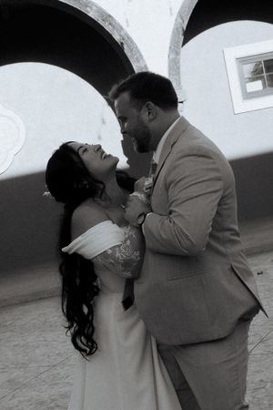 Black and white photo of a couple in wedding attire sharing a romantic moment, smiling at each other near arched walls.