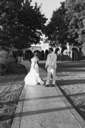Black and white photo of bride and groom walking hand in hand on garden pathway near Spanish-style arched building.