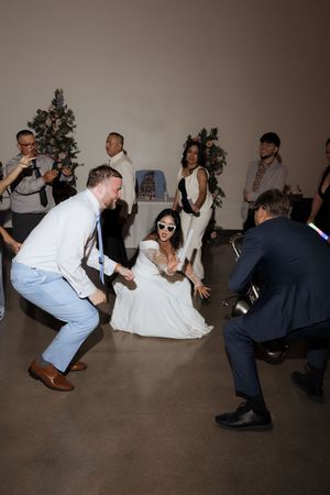 Bride in white gown and sunglasses sits on dance floor surrounded by guests at a lively wedding reception.