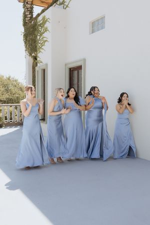 Five bridesmaids in matching dusty blue satin gowns reacting with excitement outside a white Mediterranean-style building.