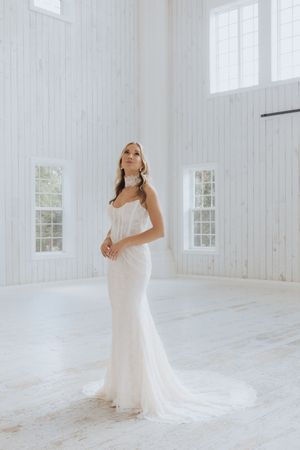 Elegant bride in a fitted white lace mermaid gown with a train stands in a bright white wood-paneled venue.